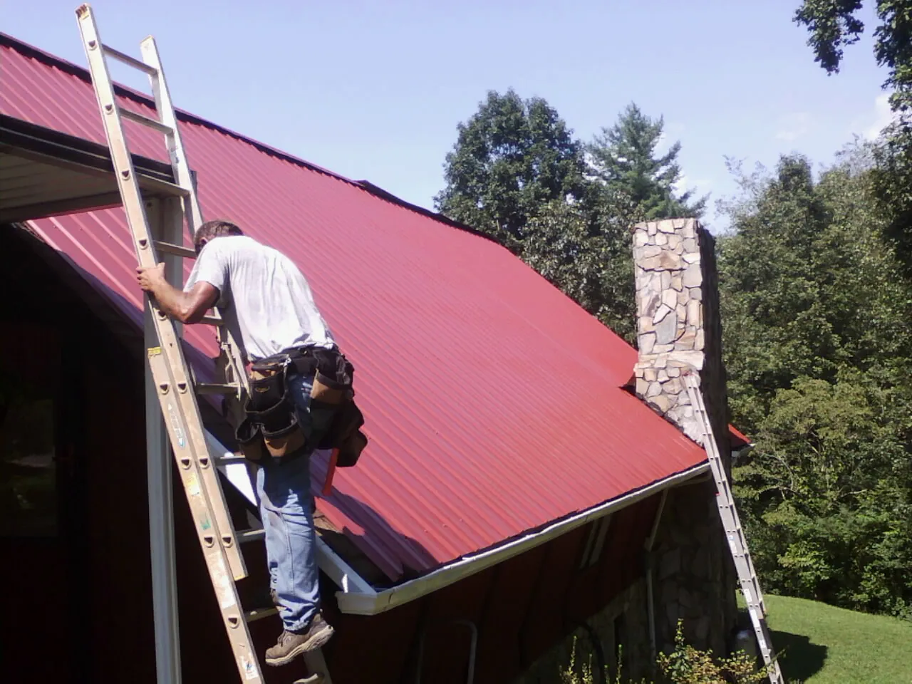 Roofer on a ladder beside a red metal roof and stone chimney — Soffit & Fascia Repair by Roofing Brothers in Palo Alto